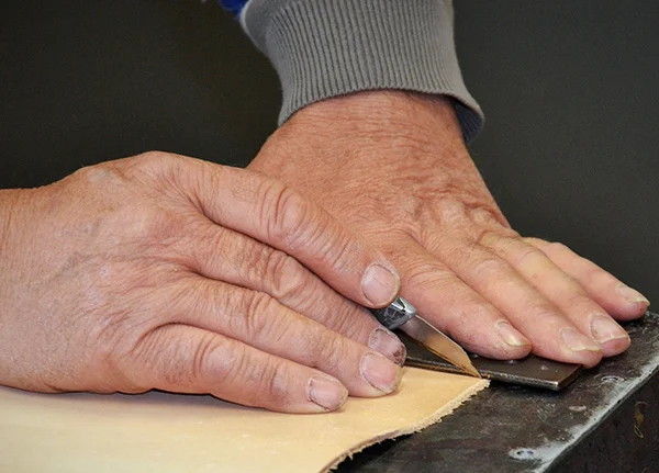 Hands holding an Exacto knife cutting through a piece of leather