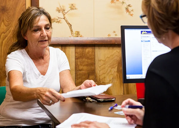 Two women sitting across from each other at an office desk and handing paperwork between them