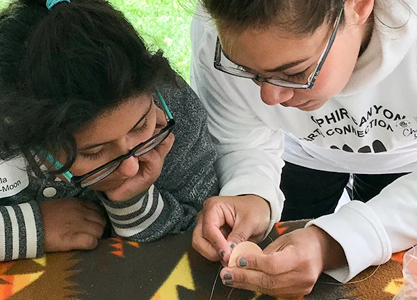 Young woman pokes a needle through a piece of fabric while a younger girl watches closely