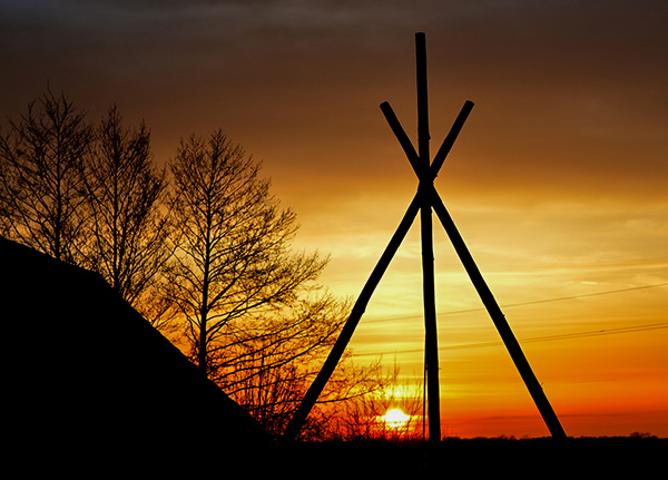 The frame of a teepee with the sun setting behind it