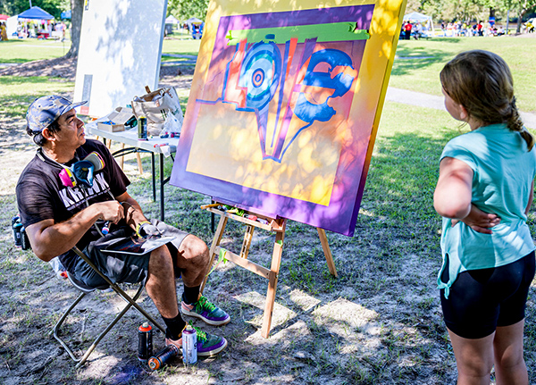 Man paints on a large canvas in a park while a young girl watches
