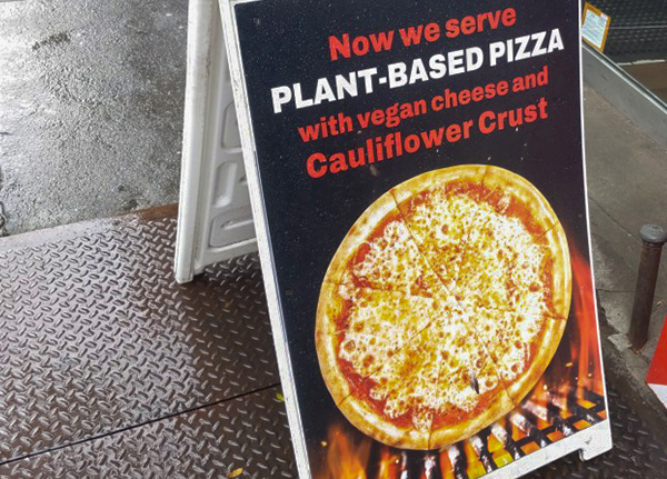 A folding sign on a sidewalk advertising that a business serves plant-based pizza with vegan cheese and cauliflower crust