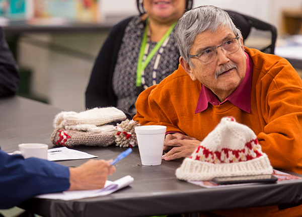 An older man sits at a table and leans in to listen to someone out of frame