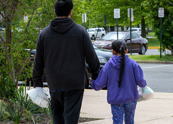 A father and a young daughter carry take out food as they walk down a sidewalk