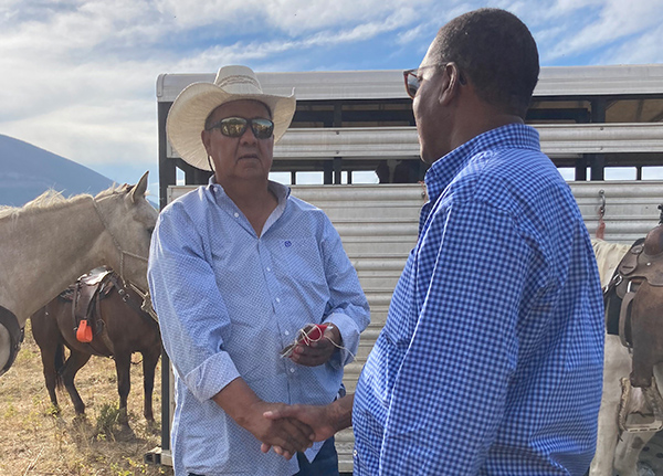 Two men shaking hands with a horse trailer and horses in the background