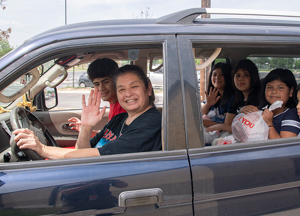 A family sitting in a car smiling and waving to the camera, one child in the back holds a to-go food bag