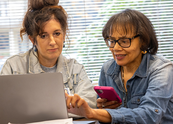 Two women sitting next to each other looking at a laptop screen, one holds a phone and points to the screen